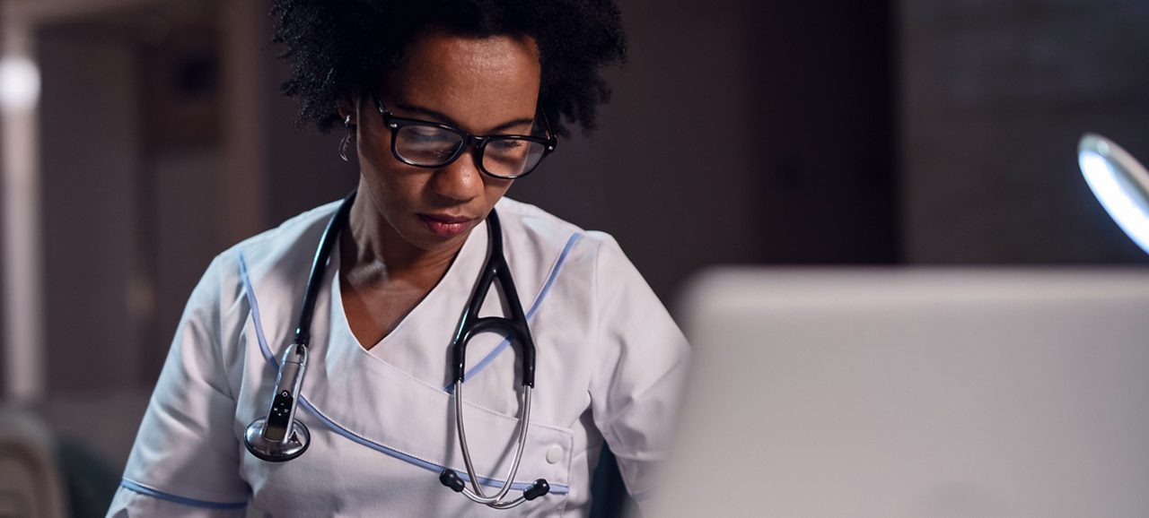 Image of a woman reading on a tablet. She is wearing glasses and a white scrub