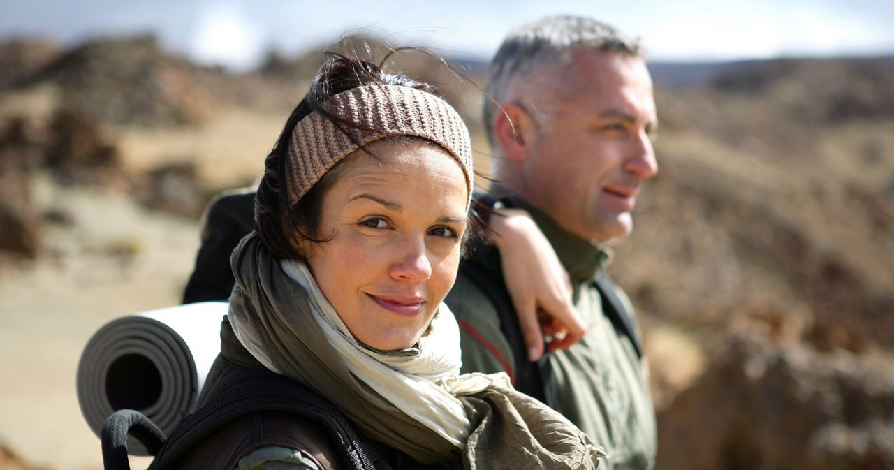Image of a woman looking at the camera. She is wearing a backpack and accompanied by a man.