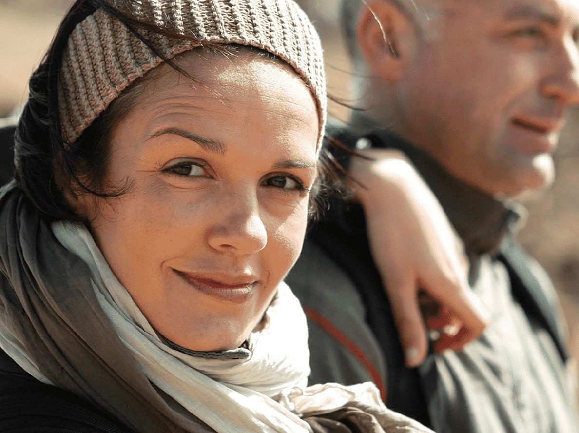 Image of a woman looking at the camera. She is wearing a backpack and accompanied by a man.