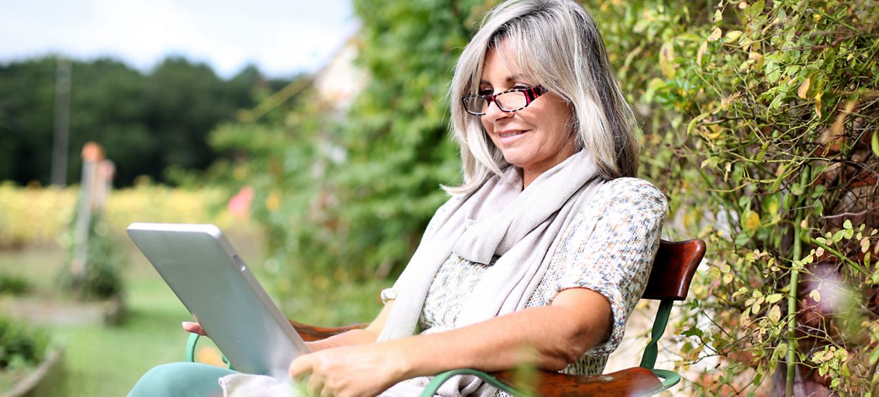 Image of a woman reading on a tablet. She is wearing glasses and a white scarf