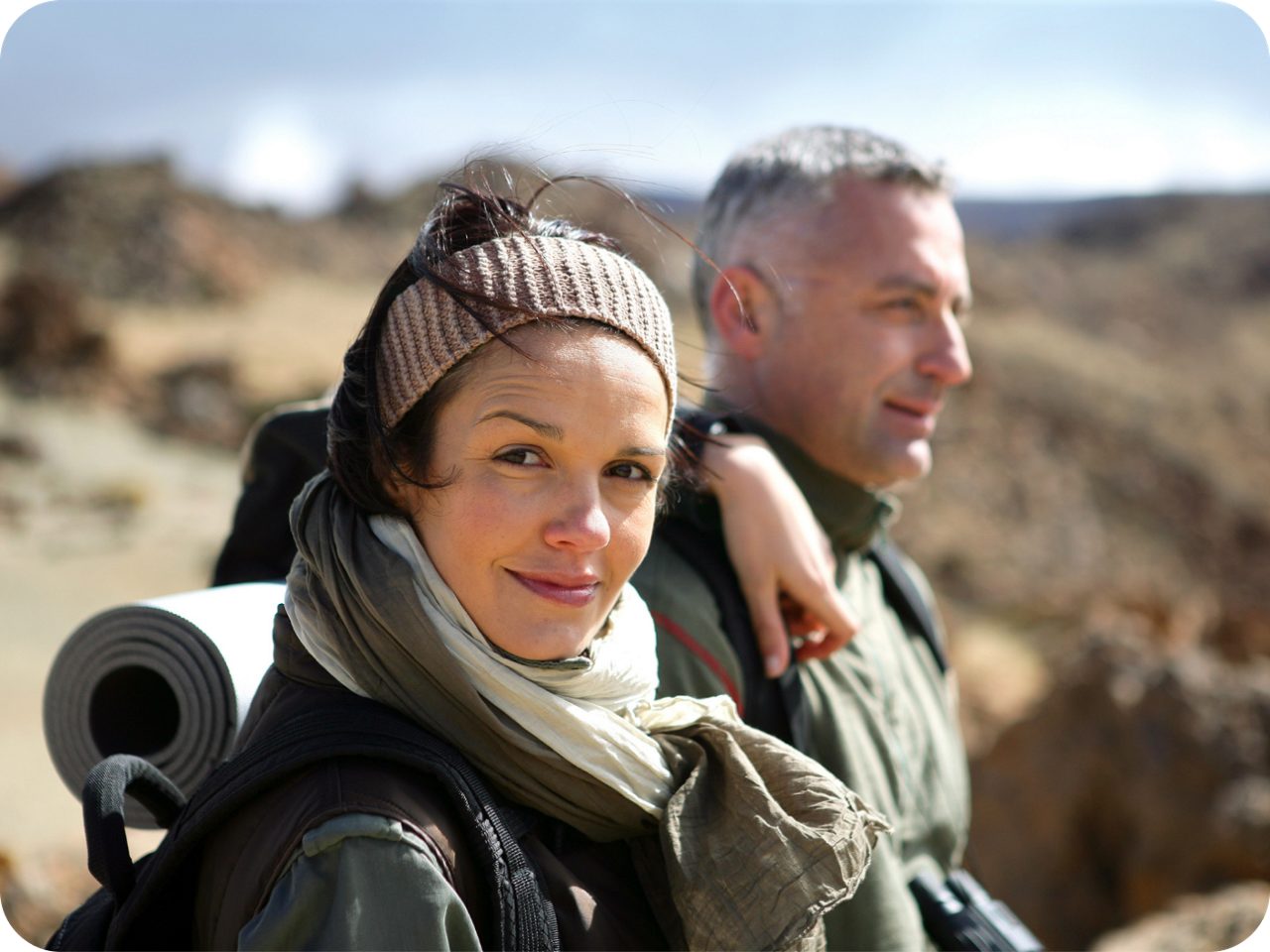 Image of a woman looking at the camera. She is wearing a backpack and accompanied by a man.