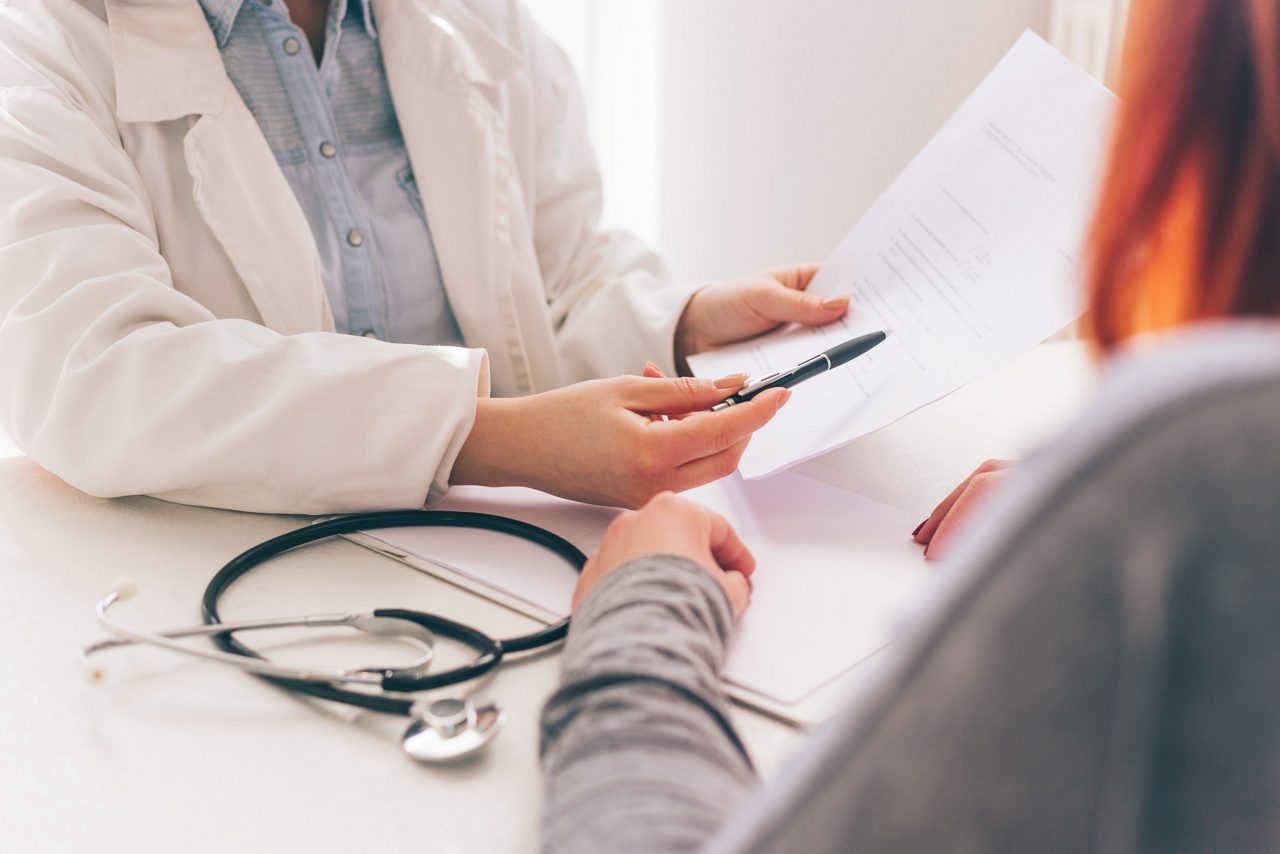 Doctor and patient are discussing about diagnosis, hands at the table. Female doctor taking notes.