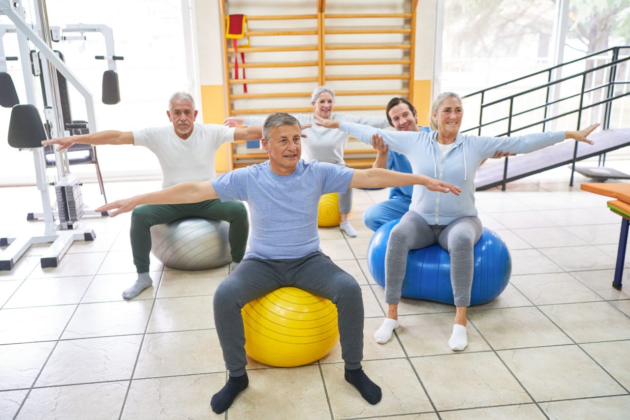 Group of elderly people exercising on fitness ball while caregiver assisting at rehabilitation center