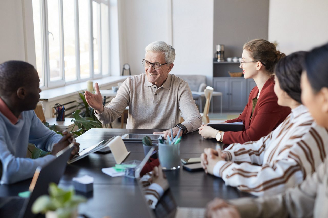 Portrait of white haired senior man instructing diverse group of people during briefing meeting in office