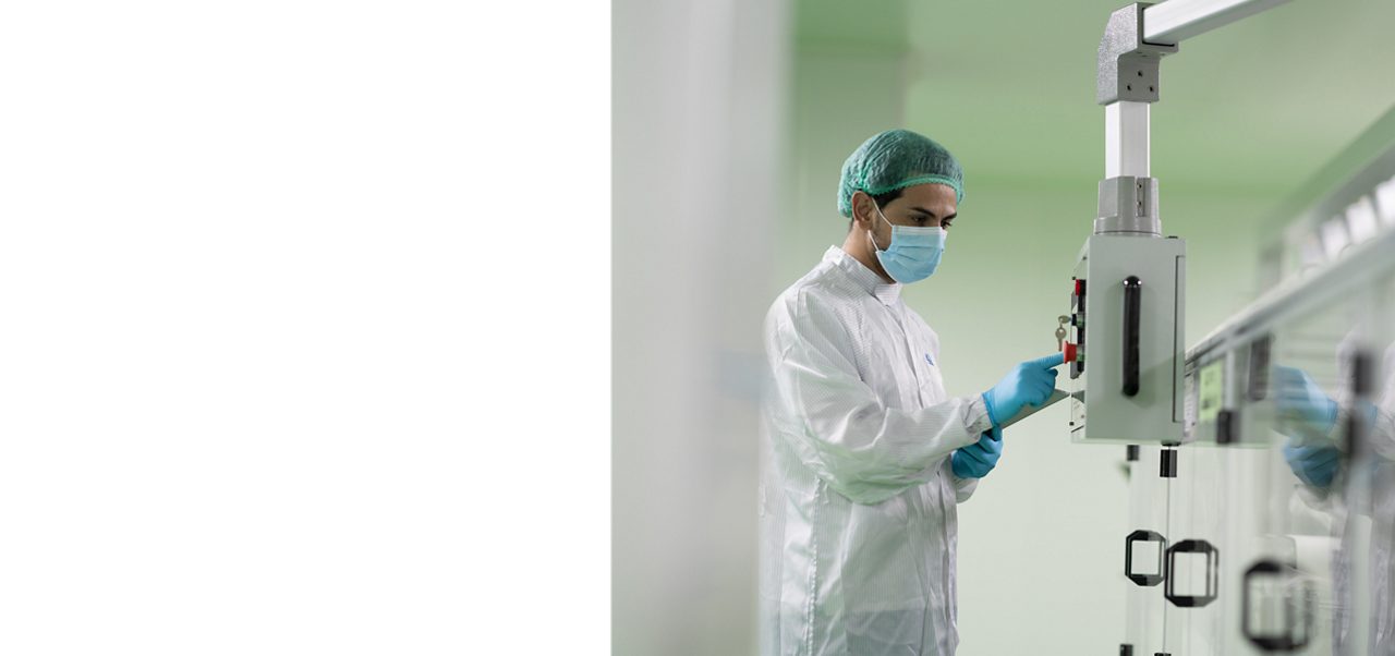 Astellas scientist wearing a white lab coat, medical mask, and rubber gloves, working with lab equipment in a high-tech research facility.